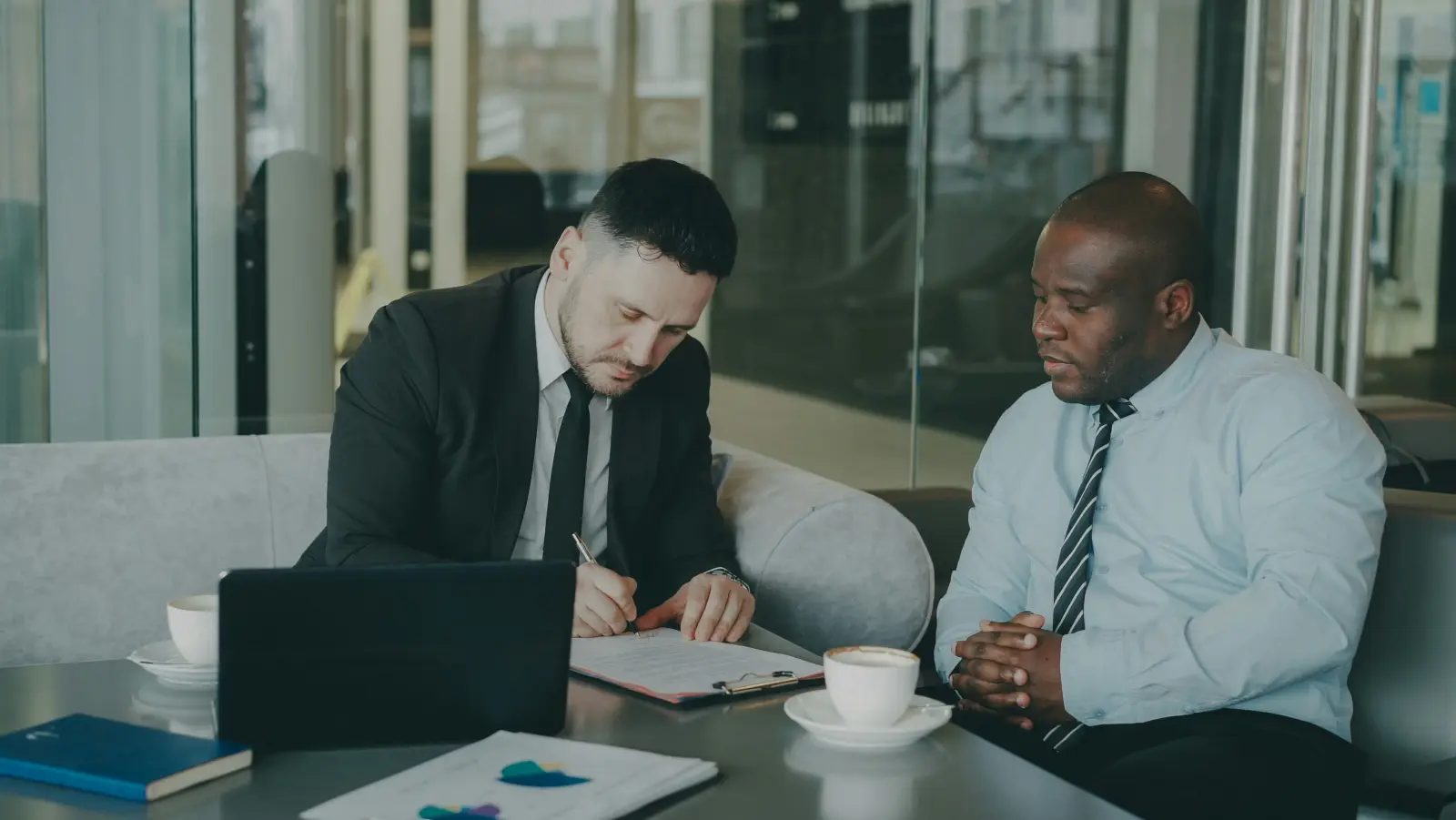Two businessmen at a table sign a partnership agreement, laptop and coffee cups nearby